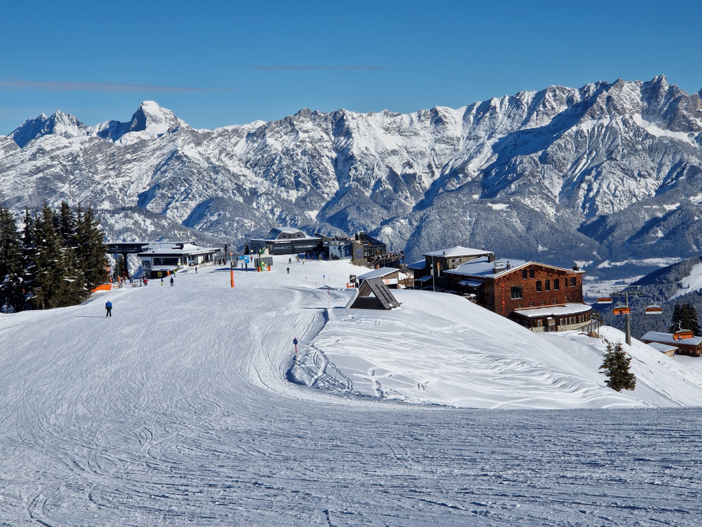 verschneite Skipiste mit Bergstationen vor winterlichem Alpenpanorama