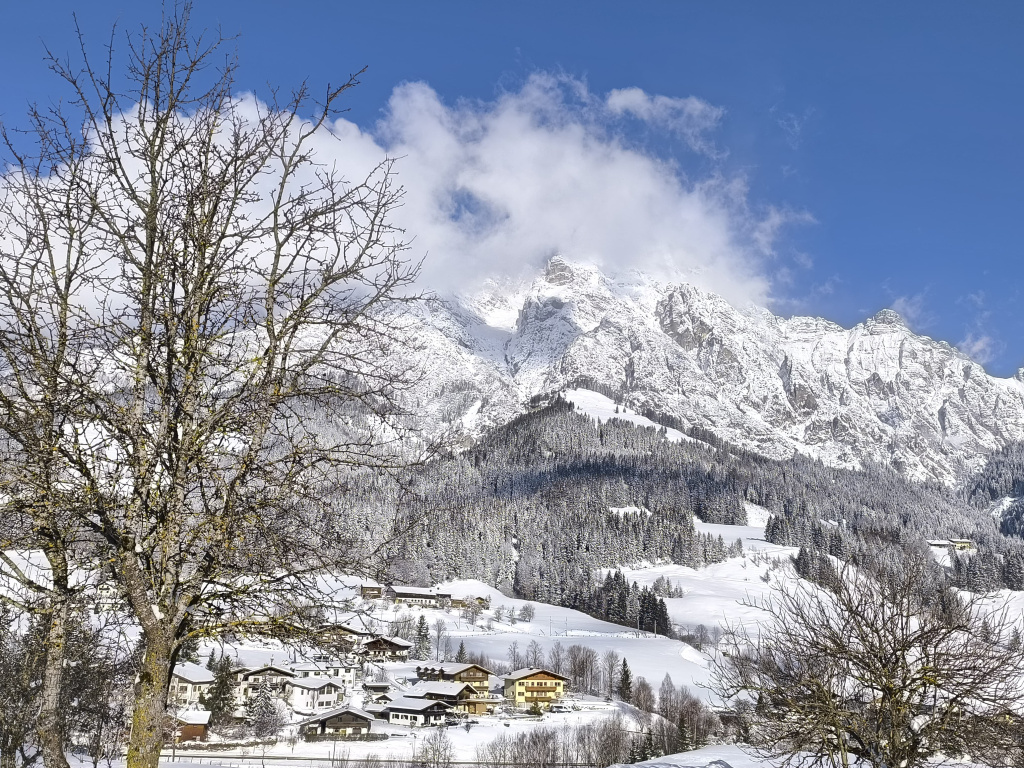 Verschneites Dorf vor schneebedecktem Gebirge unter blauem Himmel, mit kahlen Bäumen im Vordergrund